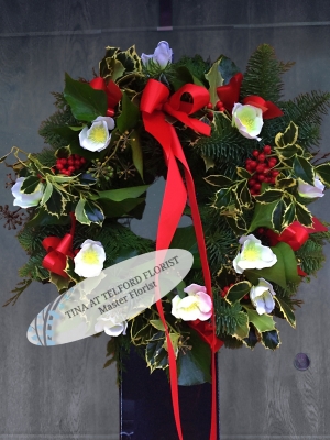 Festive wreath with red ribbons, white flowers, holly leaves, and red berries, creating a cheerful, holiday atmosphere on a wooden door.