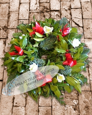 A festive wreath with holly leaves, red ribbons, and white flowers on a rustic stone background.