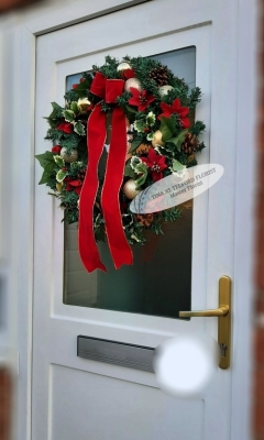 A festive wreath with red ribbons, ornaments, and greenery hangs on a white door with a frosted glass panel and golden handle, creating a cheerful holiday mood.